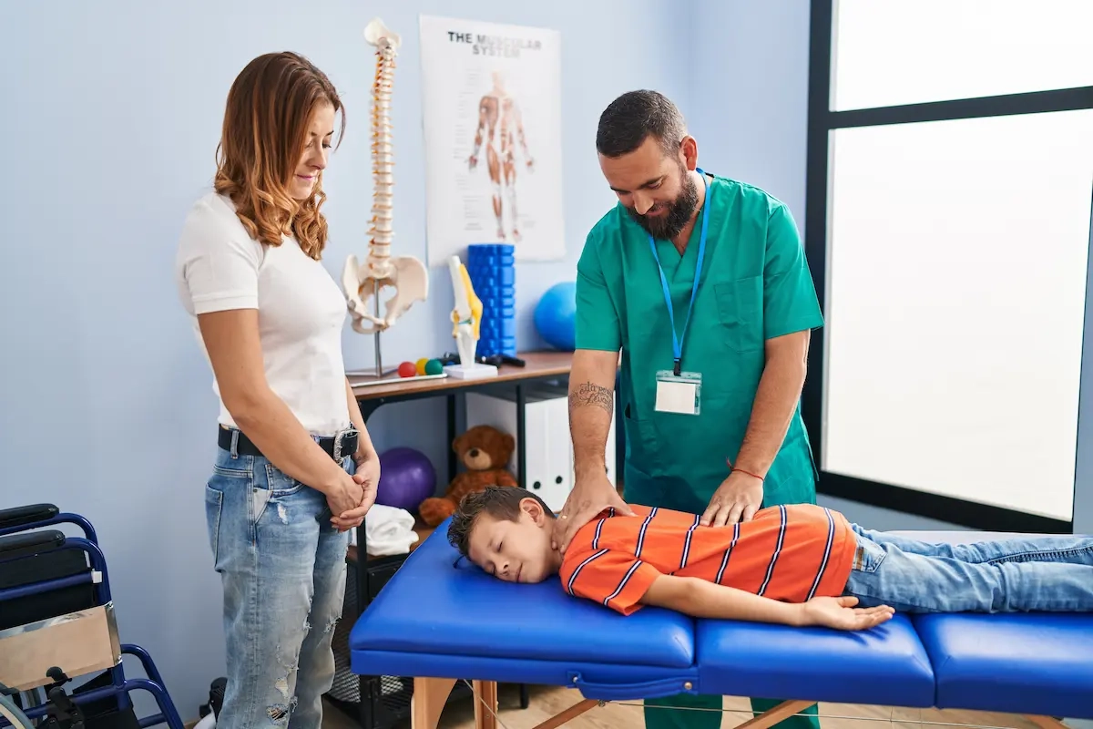 Child lying on treatment table receiving chiropractic adjustment from male chiropractor with mother watching Child receiving chiropractic adjustment from chiropractor